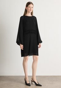 Čierne plisované šaty s dlhými rukávmi, na smockovom páse a nad kolená. Nosené s čiernymi slingback opätkami na svetlo šedém povrchu.