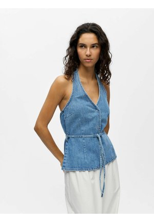 Femme aux cheveux bouclés portant un gilet en denim sans manches ceinturé et un pantalon blanc, regardant sur le côté sur un fond uni.