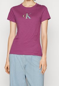 Fialové bavlněné tričko s kulatým výstřihem, krátkými rukávy a logem v pastelových barvách na prsou. Vzájemně kombino... s světle modrými kalhotami.