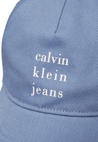 Mėlyna džinsinė kepurė su spausdintu tekstu "calvin klein jeans" balta spalva. Audinys turi tekstūruotą paviršių su matomomis siūlių detalėmis.