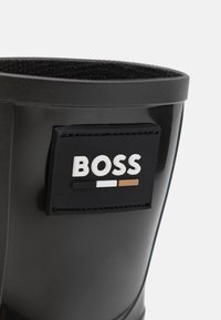 Botte en caoutchouc noire avec un patch noir cousu affichant le mot "BOSS" au-dessus de trois barres horizontales en noir, blanc et marron.