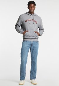 Felpa grigia con cappuccio e scritta rossa "GUESS JEANS", tasche frontali, abbinata a jeans chiari strappati e sneaker bianche.