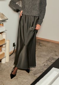 Jupe maxi en satin noir avec des plis, associée à un cardigan gris et des escarpins noirs à talons pointus. Texture lisse sur un fond neutre.