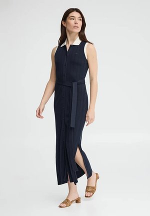 Kobieta idąca w granatowej sukience maxi na guziki bez rękawów z bocznym rozcięciem, białym kołnierzykiem, materiałowym paskiem i beżowymi sandałami na niskim obcasie.