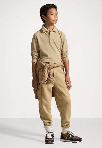 Camisa polo de mangas longas bege, calças de moletom bege, suéter de malha atado à cintura e ténis azul-marinho com detalhes amarelos. Meias brancas visíveis.