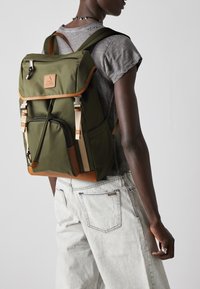 Sac à dos en nylon vert avec des accents en cuir, rabat supérieur, bretelles ajustables et poche zippée à l'avant ; porté sur une chemise grise et un jean clair.