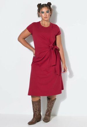 Mujer con dos moños, vestida con un vestido rojo hasta la rodilla con un nudo lateral y botas vaqueras marrones bordadas, de pie con la mano en la cadera.