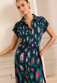 Abito camicia in blu navy con stampa animale verde e rosa, maniche corte, vita aderente e bottoni dorati lungo la parte anteriore. Tessuto dalla texture liscia.