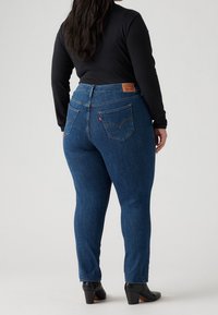 Jeans skinny de mezclilla azul con cintura alta. Cuenta con bolsillos traseros con detalle de costura y una etiqueta de marca de cuero marrón. Se llevan con botas negras.