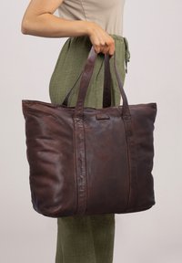 Borsa tote in pelle marrone con una texture morbida, di grande dimensione e due manici robusti, mostrata mentre viene tenuta da una persona che indossa pantaloni verdi.