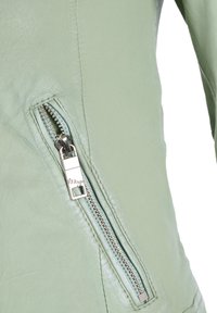 Jasnozielona skórzana kurtka z boczną kieszenią na zamek z srebrnymi elementami i teksturowanym wykończeniem. Na powierzchni widoczne są przeszycia.