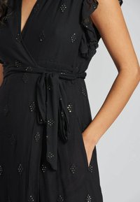 Frau in einem schwarzen Kleid mit Paillettenverzierungen, Rüschensleeves und einem Stoffgürtel, der in der Taille gebunden ist, mit einer Hand in der Tasche vor hellem Hintergrund.