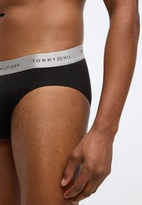 Μαύρα βαμβακερά σλιπ με ανοιχτό γκρι ελαστική ζώνη που φέρει το λογότυπο "TOMMY HILFIGER". Κοντινό πλάνο του ενδύματος φορεμένο σε μέρος του σώματος.