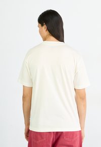 Camiseta de algodón de color crema con un corte relajado, mangas cortas y cuello redondo. Mostrada desde la parte posterior, con un detalle mínimo.