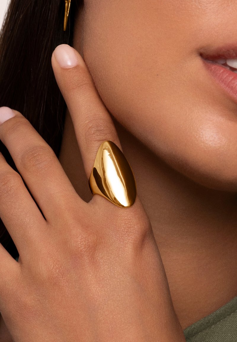 Anello in metallo dorato con una forma liscia, allungata e asimmetrica indossato su un dito. Il tono della pelle è visibile, con uno smalto per unghie rosa chiaro.