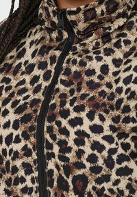 Jakna sa patentnim zatvaračem s printom, koja ima bež pozadinu sa smeškastim smeštem i crnim leopard mrljama, teksturiranu tkaninu i visoki ovratnik.