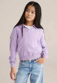 Jeune fille aux longs cheveux noirs portant un pull violet clair sur une chemise blanche et un jean bleu clair, debout avec une main dans la poche.