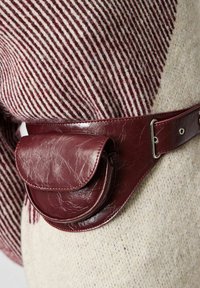 Bordový kožený pásek s kulatou, texturovanou kapsičkou, která má klopu a stříbrné kování, nošený přes světle pruhovaný svetr.