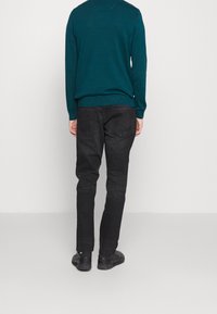 Osoba w turkusowym swetrze, czarnych dżinsach i czarnych butach stoi twarzą do gładkiej białej ściany, z rękami przy bokach.