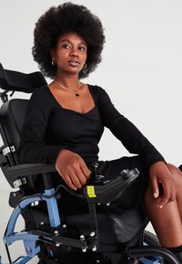 Αυτοπεποιθητική γυναίκα με αφρικανικό χτένισμα, καθισμένη σε μια μαύρη και μπλε ηλεκτρική αναπηρική καρέκλα, ντυμένη με ένα μαύρο φόρεμα και χρυσά κοσμήματα.