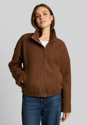 Woman with brown textured zip-up jacket and blue jeans stands against plain light background, hands in pockets, looking slightly up and smiling.