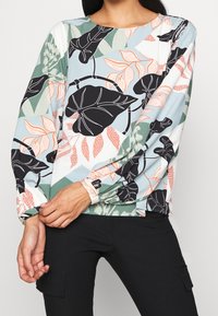 Camicia a maniche lunghe con motivo floreale in azzurro chiaro, caratterizzata da foglie nere e fiori pesco. Tessuto morbido con una vestibilità rilassata e maniche arricciate.