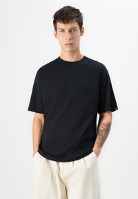 Jovem com cabelo curto e escuro, vestindo uma t-shirt preta e calças beige claras, com as mãos nos bolsos, mostrando uma tatuagem no antebraço esquerdo.