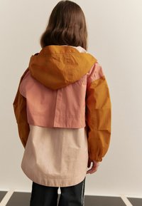 Färgblockerad huva-jacka med orange, rosa och beige paneler, tillverkad av lätt material med en avslappnad passform och texturerad yta.