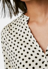 Hvid bluse med sorte prikker, der har en krave og knapdetaljer. Stoffet ser letvægtsagtigt ud med en glat tekstur.
