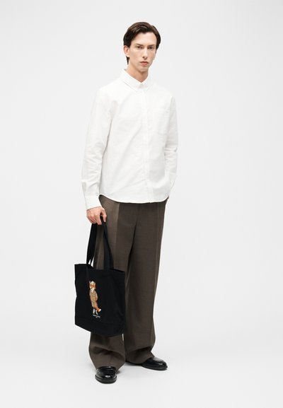 Άνδρας που φοράει λευκό πουκάμισο με κουμπιά και καφέ παντελόνι, κρατώντας μαύρη τσάντα tote με εικονογράφηση αλεπούς, στέκεται σε λευκό φόντο.