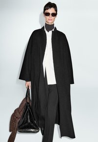 Persona che indossa un cappotto oversize nero, una camicia bianca con bottoni, pantaloni neri, occhiali da sole, mentre porta una grande borsa nera e un capo lavorato a maglia marrone.