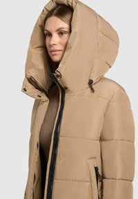 Beige polsterdatud jope suure kapuutsiga, millel on nuppe ja eesmine lukku, mustad aktsendid lukudel ja küljetaskutega.