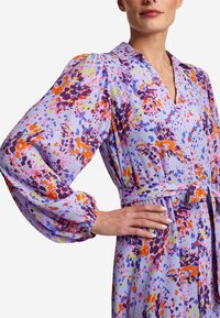Fioletowa sukienka w kwiatowy wzór z długimi bufkami, kołnierzykiem i paskiem w talii. Materiał wygląda na gładki, z jasnymi plamami pomarańczowego i zielonego koloru.
