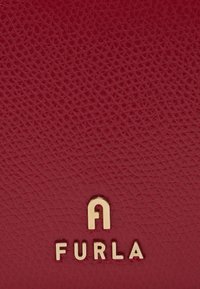 Czerwona skórzana tekstura z widocznym usłojeniem. Złote okucia z literą "A" oraz nazwą marki "FURLA" wytłoczonymi w wyraźnej czcionce.