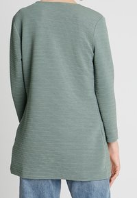 Långärmad topp i texturerat, dämpat grönt tyg. Har en rund halsringning och en avslappnad passform, med horisontell ribbning över hela.