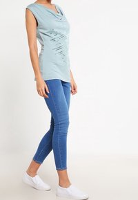 Top sin mangas azul claro con escote drapeado y diseño plateado impreso; combinado con jeans ajustados azules y zapatillas blancas sin cordones.