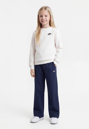 Ung pige med langt blondt hår iført en hvid Nike-sweatshirt, marineblå joggingbukser og hvide sneakers, stående og smilende.