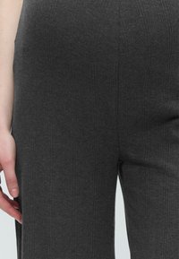 Κοντινό πλάνο ενός ατόμου που φοράει σκούρο γκρι πλεκτό παντελόνι με ρίγα, με το αριστερό χέρι χαλαρό δίπλα στο σώμα, σε απλό φόντο.