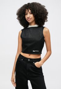 Žena kučeravých vlasov v čiernom bezrukávovom krátkom topu a čiernych džínsach s vysokým pásom, stojaca s jednou rukou v vrecku na jednoliatom pozadí.