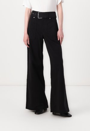 Persona vistiendo pantalones negros de talle alto y pierna ancha con un cinturón grande, combinados con zapatos negros de tacón y una camiseta blanca de manga corta.