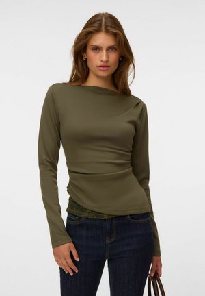 Femme portant un haut à manches longues vert olive et un jean bleu foncé, debout, une main tenant la sangle d’un sac marron sur un fond clair uni.