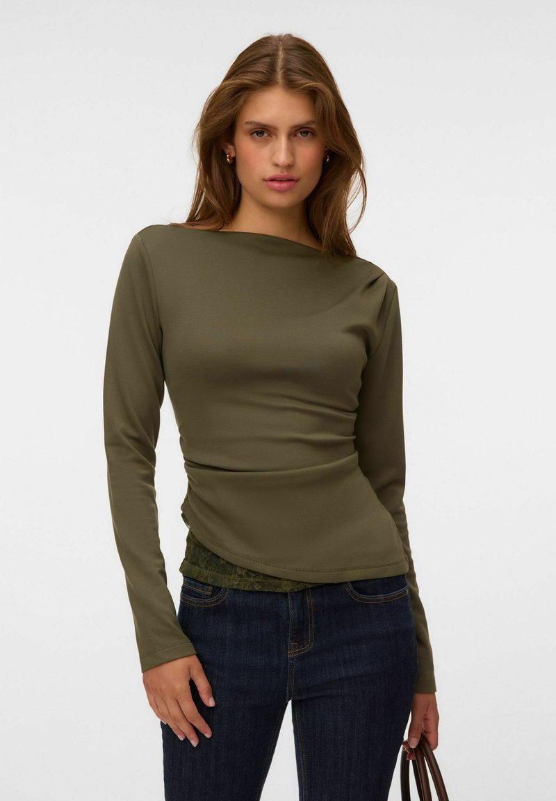 Femme portant un haut à manches longues vert olive et un jean bleu foncé, debout, une main tenant la sangle d’un sac marron sur un fond clair uni.