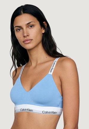 Donna con capelli lunghi e scuri che indossa un bralette Calvin Klein azzurro chiaro con spalline elastiche bianche con logo e banda.