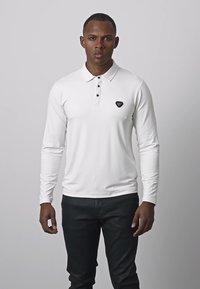 Polo de manga larga blanco con tres botones negros y un pequeño logo negro en el pecho izquierdo, combinado con pantalones negros. Textura lisa.