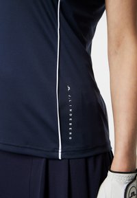 Marinblå performance-shirt med slim fit, vita sidoseams och en logotyp. Ackompanjeras av en plisserad marinblå kjol och en vit golfhandske.