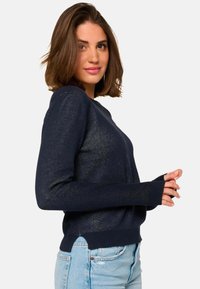 Pull en maille texturée bleu marine à manches longues, col rond, avec ouvertures pour les pouces et ourlet côtelé. Associé à un jean taille haute bleu clair.