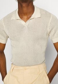 Krótka koszulka polo z krótkim rękawem w kolorze kremowym, z dekoltem w serek, wykonana z otwartym wzorem dzianiny i dopasowanym krojem. Noszona z jasnobeżowymi spodniami.