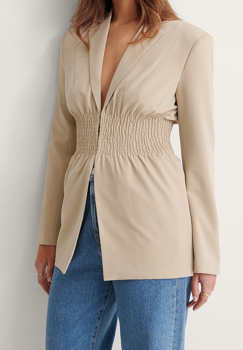 Femme portant un blazer beige avec un détail de taille élastique froncée, associée à un jean bleu, debout contre un mur clair.
