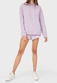 Mujer vestida con una sudadera ligera color púrpura y pantalones cortos a juego, llevando zapatillas blancas, de pie frente a un fondo blanco liso.