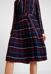 Kobieta ubrana w plisowaną sukienkę w granatową, czerwoną i niebieską kratę, z długimi rękawami oraz dopasowanym paskiem w paski w talii.